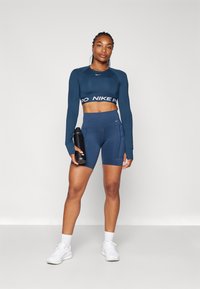 Tengerészkék, hosszú ujjú crop top fehér logós pánttal, magas derekú tengerészkék sortokkal, zsebekkel, valamint fehér sportcipőkkel. Fekete water bottle-t tart a kezében.