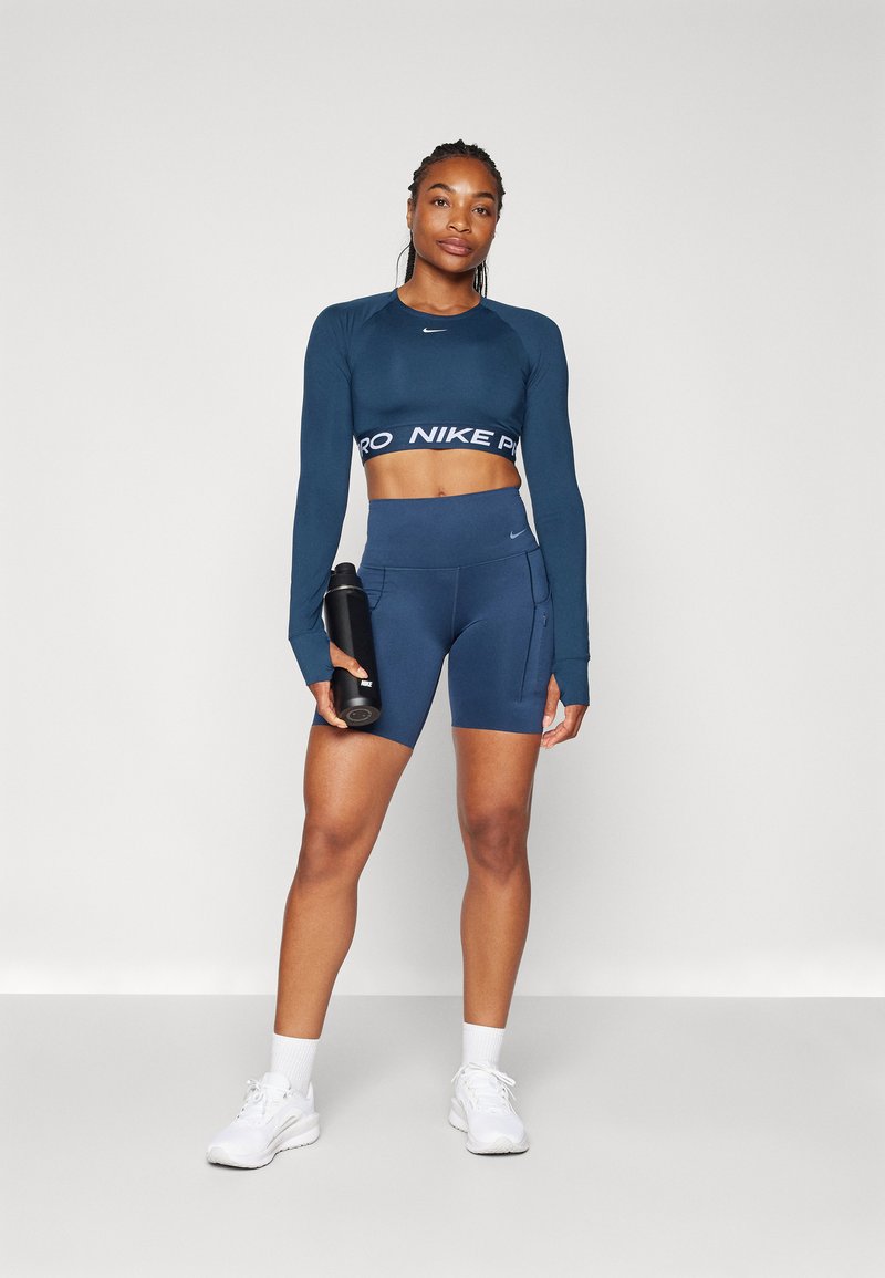 Marineblaues Langarm-Crop-Top mit einem weißen Logoband, kombiniert mit hochtaillierten marineblauen Shorts mit Taschen und weißen Turnschuhen. Hält eine schwarze Wasserflasche.