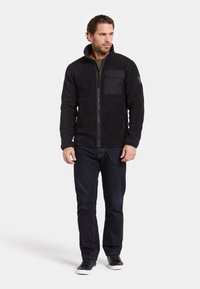 Mustavilla fleece-takilla, jossa on korkea kaulus, etuvetoketju ja rintatasku. Yhdistetty tummiin farkkuihin ja mustiin kenkiin. Rakennekerroksinen kangas.