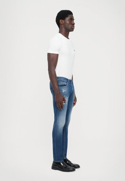 T-shirt bianco a maniche corte, jeans in denim blu con dettagli invecchiati e scarpe nere, mostrati in profilo laterale su uno sfondo semplice.