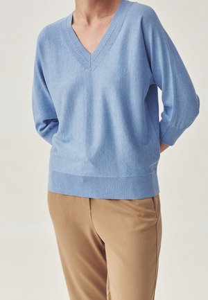 Personne portant un pull bleu clair à col en V avec des manches trois-quarts et un pantalon beige, debout les mains dans le dos.