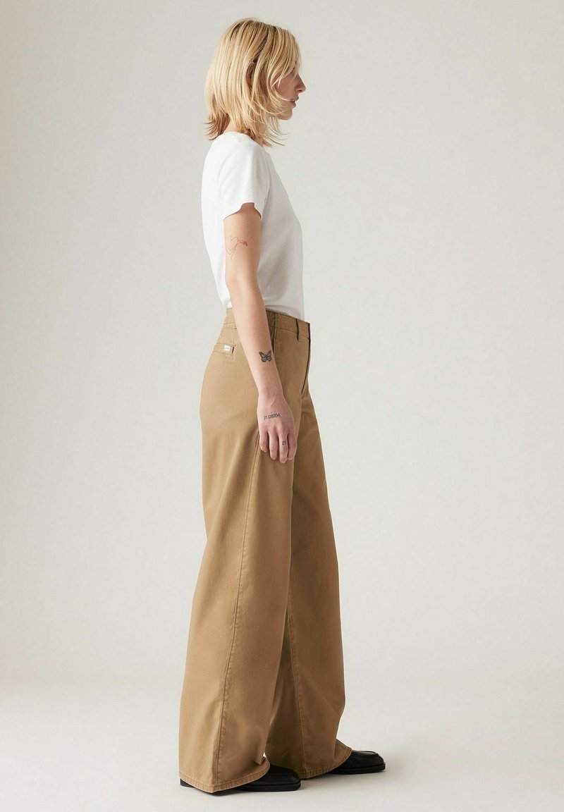 Pantaloni larghi color khaki con vita alta, realizzati in un tessuto robusto, abbinati a una t-shirt aderente bianca. Linee pulite e senza motivi visibili.