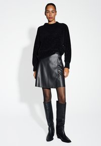 Schwarzer Strickpullover mit langen Ärmeln, kombiniert mit einem schwarzen Lederwickelrock und kniehohen Lederstiefeln, was einen monochromatischen Look schafft.
