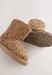 Bottes en daim marron pour tout-petits avec motifs de visages de lapin noirs et semelles en caoutchouc texturées, montrées de côté et de dessous sur fond blanc.