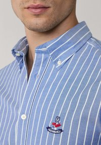 Camisa de cuadros azul y blanca con cuello rígido, que presenta un pequeño logo bordado en rojo y azul cerca del bolsillo del pecho.