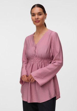 Frau trägt eine langärmelige, locker sitzende rosa Bluse mit geraffter Taille und schwarze Hose, steht vor einem schlichten hellen Hintergrund.