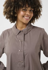 Rudas mygtukų marškiniai su plačiu apykakle, turintys plokštelę ir subtilų blizgesį. Lygi tekstūra, papildyta apvaliais, šviesių spalvų mygtukais.