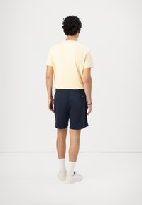 Pantaloni scurți bleumarin cu talie elastică, asortați cu un tricou galben deschis. Ținuta este completată cu șosete albe și adidași de culoare deschisă.