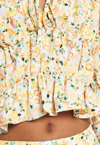 Top peplum floral com fundo branco, apresentando padrões amarelos, laranjas e verdes, decote com cordão e barra com babados.