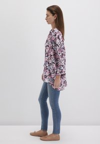 Blumenmuster-Tunika in Rosa- und Lilatönen, mit lockerer Passform und langen Ärmeln. Getragen mit blauen Skinny-Jeans und braunen flachen Schuhen.