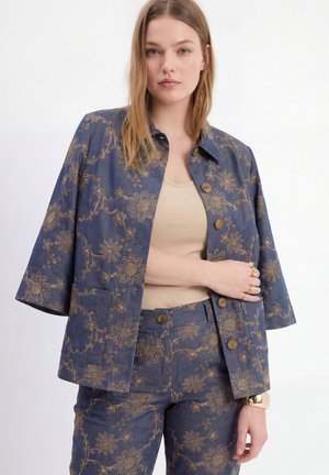 Jonge vrouw met steil haar draagt een blauwe jas met bloemenpatroon en bijpassende broek over een beige top, poserend tegen een witte achtergrond.