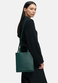 Zielona skórzana torba typu tote z dwoma krótkimi uchwytami i regulowanym paskiem na ramię. Torba charakteryzuje się gładką fakturą i minimalistycznym designem.