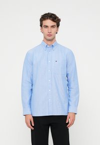 Jasnoniebieska koszula zapinana na guziki z pionowymi białymi paskami, z jedną kieszenią na piersi i kołnierzykiem zapinanym na guziki, wykonana z gładkiego materiału.