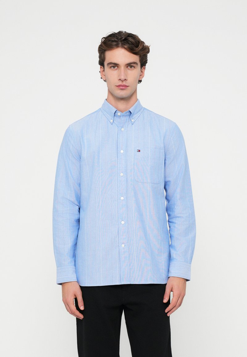 Jasnoniebieska koszula zapinana na guziki z pionowymi białymi paskami, z jedną kieszenią na piersi i kołnierzykiem zapinanym na guziki, wykonana z gładkiego materiału.