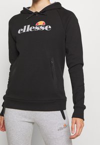 Czarna bluza z kapturem z dużym białym logo „ellesse”, kieszenią kangurką i sznurkiem do regulacji kaptura. Materiał wydaje się miękki, a krój dopasowany.