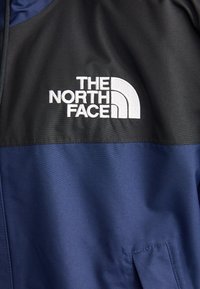 Marineblå jakke med en sort overdel, lynlås i lukning, og det hvide broderede 'The North Face' logo på brystet.