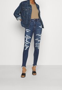 Jeansová bunda s ohrnutými rukávy přes béžový top, spojená s tmavě modrými roztrhanými skinny džíny a černými skládacími sandály, s třepenými okraji.