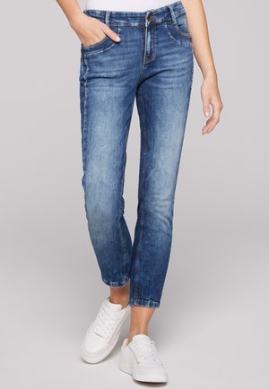 Jeans de denim azul con un acabado ligeramente desgastado, diseño de cinco bolsillos y corte ajustado, combinados con zapatillas blancas.