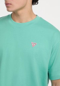 Helles grünes T-Shirt mit kurzen Ärmeln aus Baumwolle, mit einem roten, dreieckigen Logo-Patch auf der Brust, das den Text "GUESS" enthält. Glatte Textur.