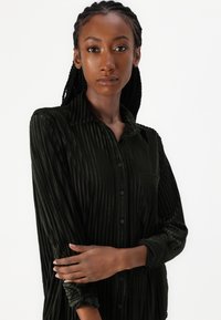 Camisa às riscas verde escuro, de tecido suave. Apresenta colarinho, abertura com botões à frente e um único bolso no peito. Mangas longas com punhos enrolados.