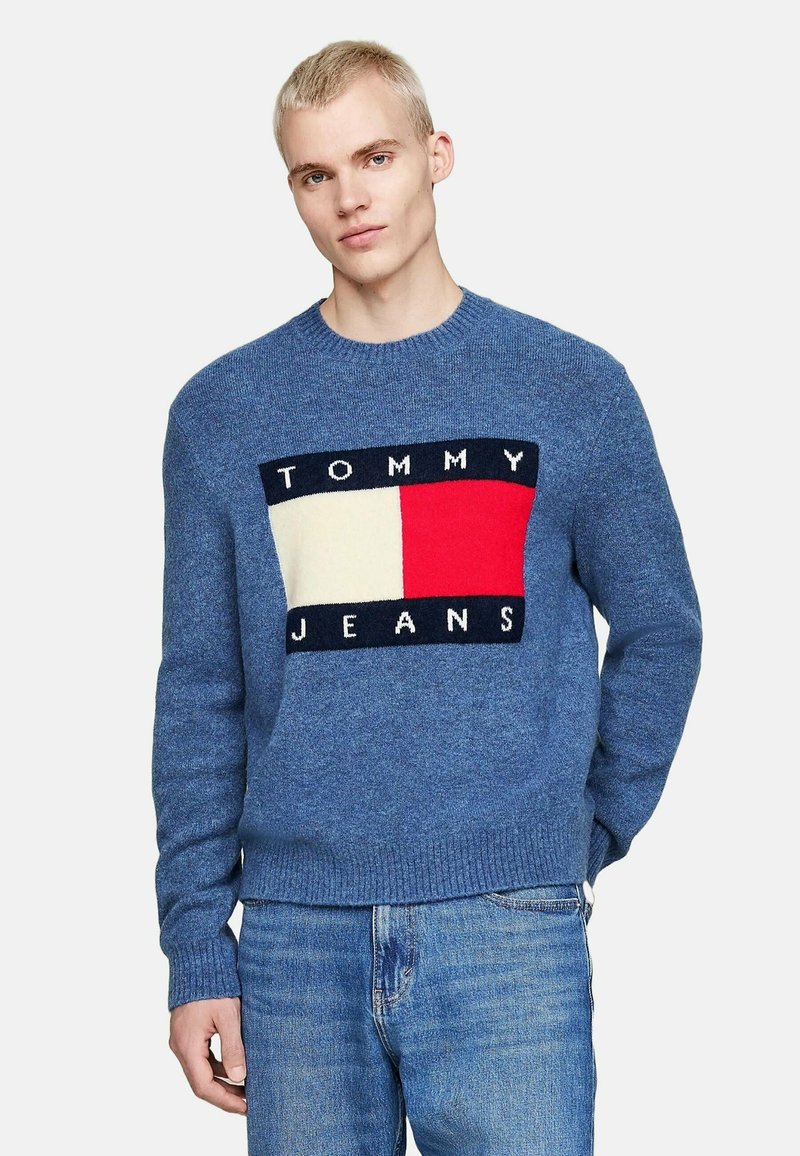 Suéter azul de punto con cuello redondo que presenta un parche gráfico de "TOMMY JEANS" en varios colores y puños y dobladillo acanalados en contraste.