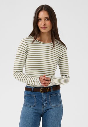 Jeune femme portant une chemise à manches longues rayée vert et blanc, jean bleu et ceinture marron, debout les mains jointes.