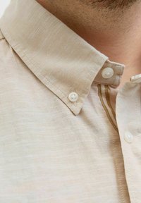 Chemise beige à col, en tissu texturé, avec deux boutons blancs et un détail à rayures verticales le long de la patte de boutonnage.