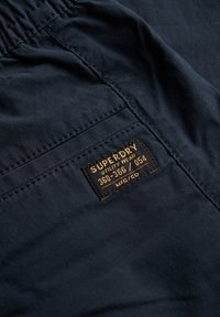 Tissu bleu marine à texture lisse avec une étiquette noire carrée affichant "SUPERDRY" et des détails sur les vêtements utilitaires.
