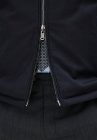 Marinblå zip-up jacka med silverfärgad detalj, som avslöjar ett mönstrat slips och en ljusblå skjorta. Svart randig byxa syns i botten.