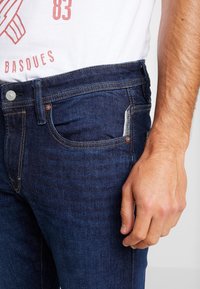 Jeans de mezclilla azul oscuro con un ajuste slim. Las características incluyen costuras en contraste, un bolsillo frontal y una pequeña etiqueta tejida en el costado.