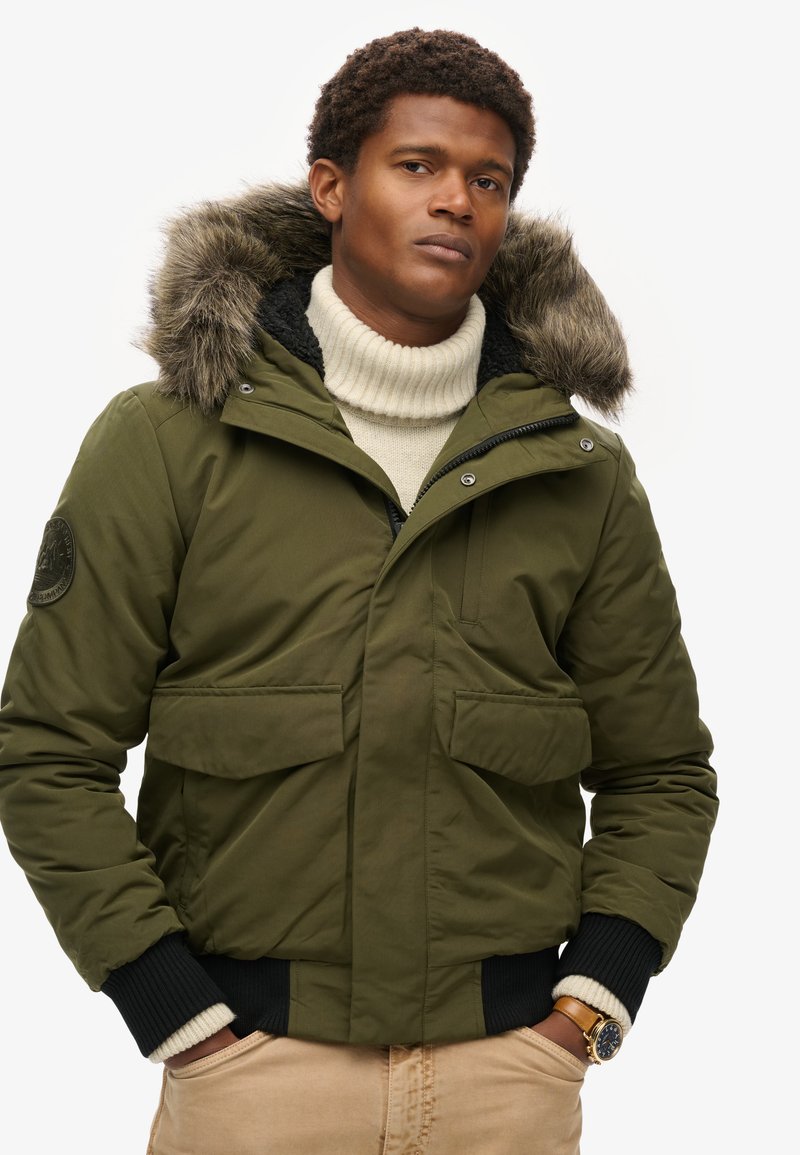 Uomo con giacca invernale verde con cappuccio in pelliccia, maglione a collo alto crema, pantaloni beige e orologio da polso, mani nelle tasche, espressione neutra.
