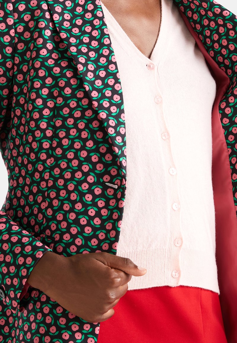Personne à la peau foncée portant un cardigan rose boutonné sous une veste noire à motif floral rose, tenant la veste ouverte pour montrer la doublure.