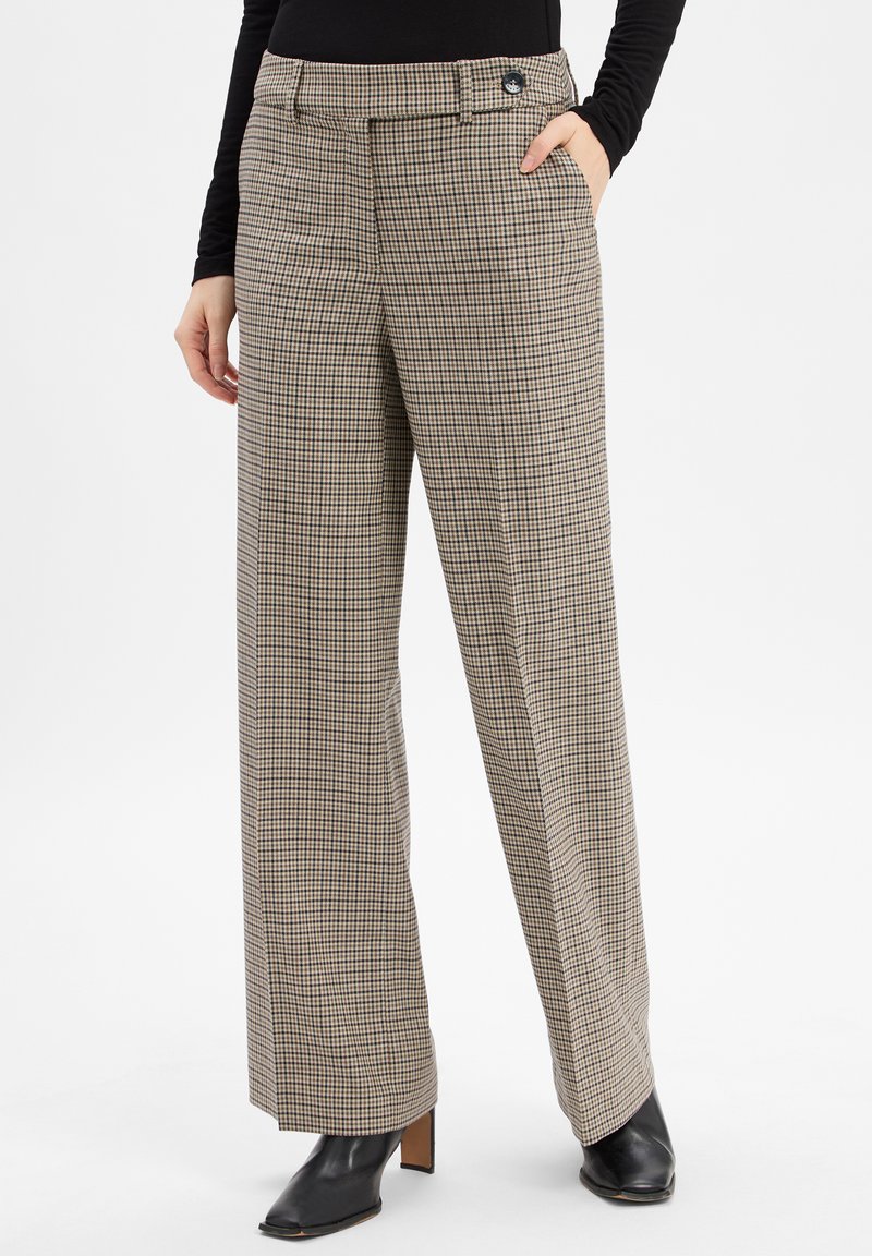 Frau, die eine beige und schwarze kariierte weit geschnittene Hose mit Seitentaschen trägt, kombiniert mit schwarzen Absatzstiefeletten und einem schwarzen Langarmoberteil.