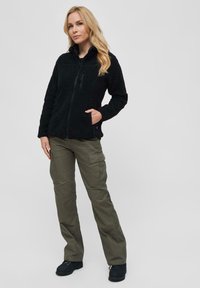 Zwarte fleece jas met een hoge kraag, ritssluiting aan de voorkant en twee zijzakken, gecombineerd met olijfgroene cargo-broeken en zwarte laarzen.