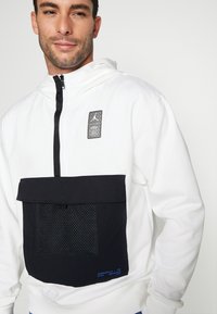 Hvid hættejakke med sorte detaljer og hætte, der har en mesh lomme og logo patch. Teksten inkluderer blå detaljer nederst. Blødt, let materiale.