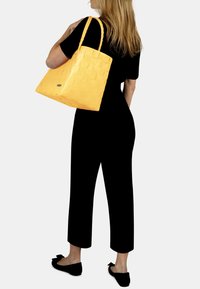 Gelbe geflochtene Tragetasche mit strukturiertem Finish und geflochtenen Griffen, ausgestattet mit einem kleinen Metall-Logodetail. Modell trägt ein schwarzes Outfit.