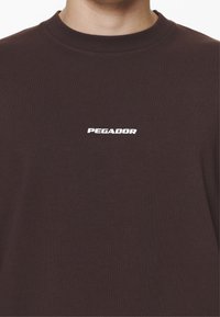 Közeli kép egy sötétbarna, kerek nyakú pólót viselő személyről, amelyen fehér "PEGADOR" felirat található, középen, a mellkason.
