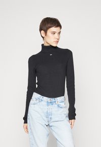 Maglione a collo alto nero a coste con maniche lunghe, design aderente e un piccolo logo nella zona del collo; abbinato a jeans chiari a vita alta.