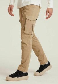 Beżowe spodnie cargo wykonane z bawełny, z wieloma bocznymi kieszeniami, w luźnym kroju, w zestawie z czarnymi sneakersami na grubej podeszwie.