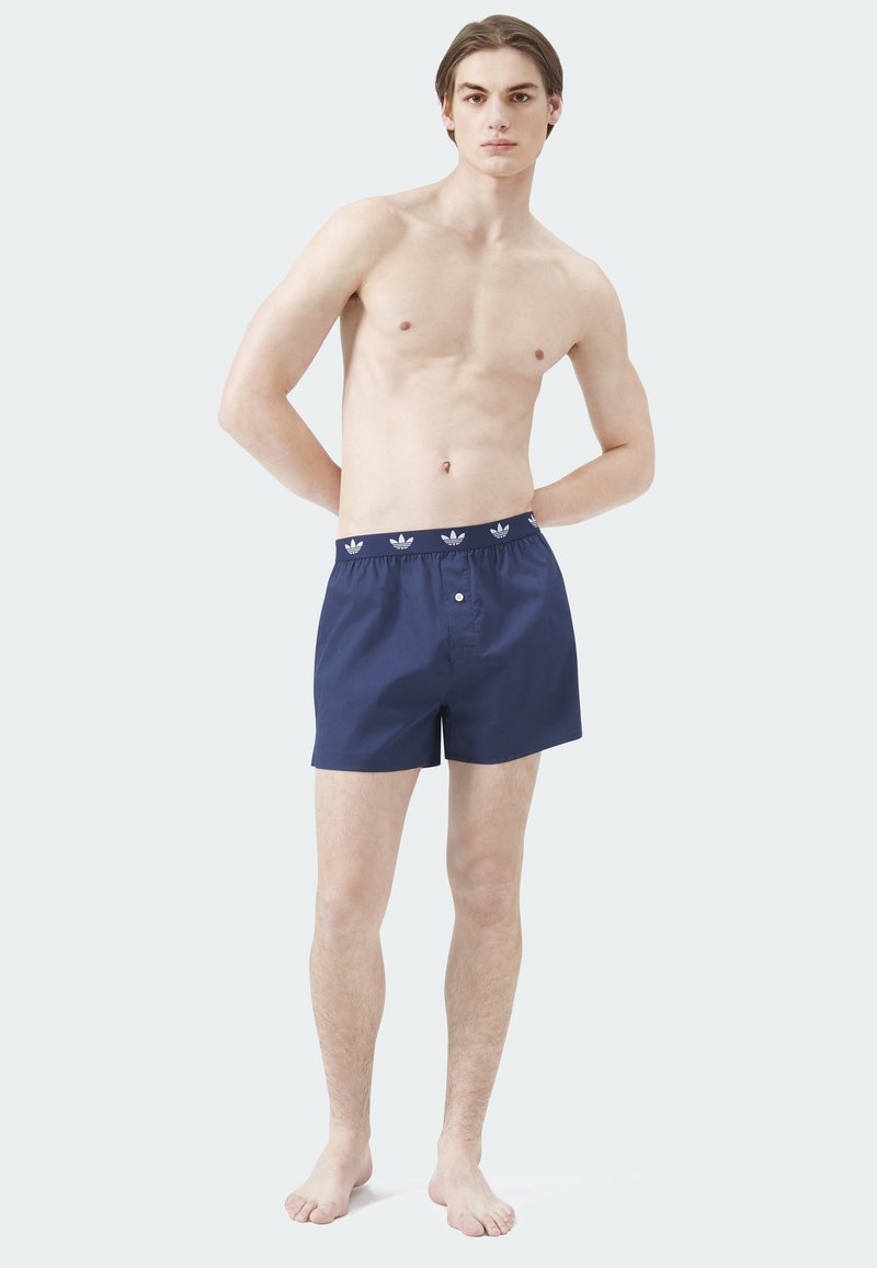 Marineblaue Badeshorts mit elastischem Bund und weißen Logo-Akzenten. Glatte Textur, Knopfverschluss und mittellange Beinlänge bis zur Mitte des Oberschenkels.