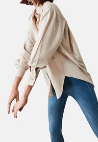 Donna che indossa una camicia beige oversize con bottoni e maniche arrotolate, abbinata a jeans skinny blu, mentre posa con le braccia tese in avanti.