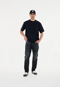 Črna kratka majica s kratkimi rokavi, temno modri jeans z obrabo in bele superge z mornarsko modrimi detajli, v kombinaciji z črno kapo z belim logotipom.