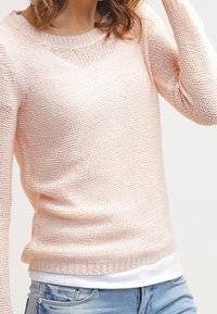 Pull rose clair en tricot avec un col rond, tissu texturé, manches longues et ourlet côtelé, superposé à une chemise blanche.
