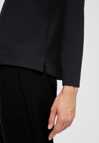 Zwarte textuur top met een honingraatpatroon en lange mouwen, gecombineerd met zwarte broeken. De zoom heeft zijsplitten voor extra detail.