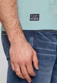 Svetlo modra bombažna majica z črno etiketom, ki prikazuje koordinate in ime blagovne znamke, v kombinaciji z temno modrimi traper hlačami, ki poudarjajo šive.