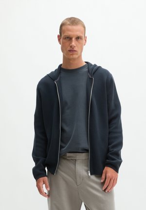 Ein junger Mann mit kurzen Haaren trägt einen dunklen Kapuzenpullover mit Reißverschluss über einem dunklen T-Shirt und hellen Hosen vor einem einfarbigen weißen Hintergrund.