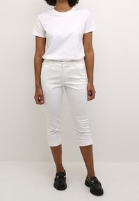 T-shirt blanc à manches courtes associée à un pantalon blanc ajusté et court. Le modèle porte des mocassins noirs avec un accent doré. Tissu lisse et coupe ajustée.