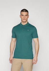 Tirkizna polo majica od pamuka s ovratnikom, plavo-bijelom gumbima, i suptilnim brendiranim logotipom na lijevoj strani prsnog koša.