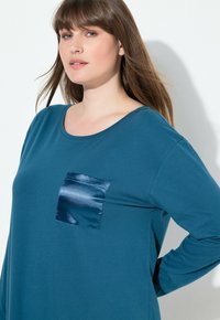 Camisa de manga larga en color verde azulado, hecha de una tela suave, con un bolsillo de parche de satén azul marino brillante en el lado izquierdo del pecho. Cuello redondeado y corte holgado.