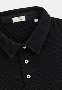 Polo noir à côtes avec un col structuré, patte de boutonnage à cinq boutons et une poche poitrine simple. L'étiquette indique la taille L, détails des boutons blancs.
